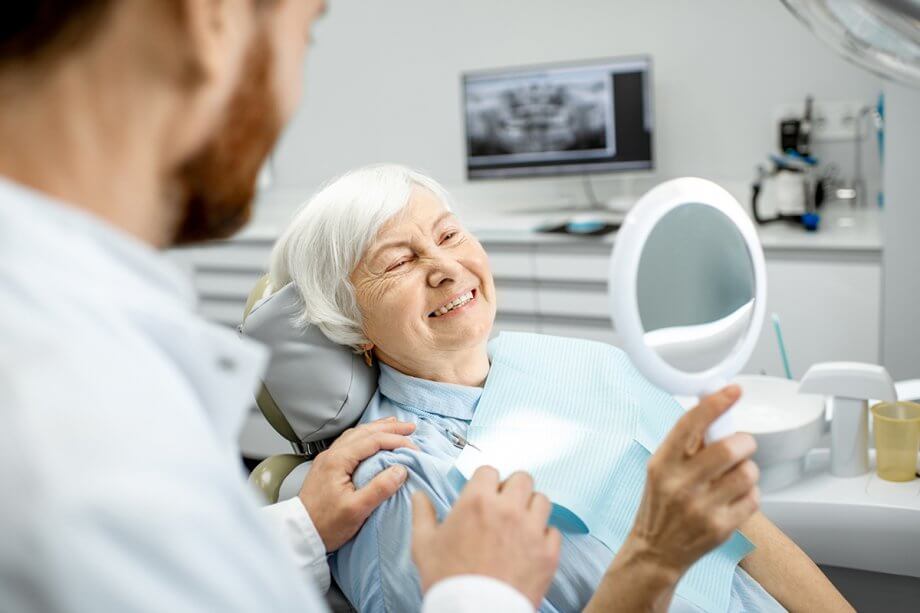 mature woman in dental chair smiles at herself in a hand mirror