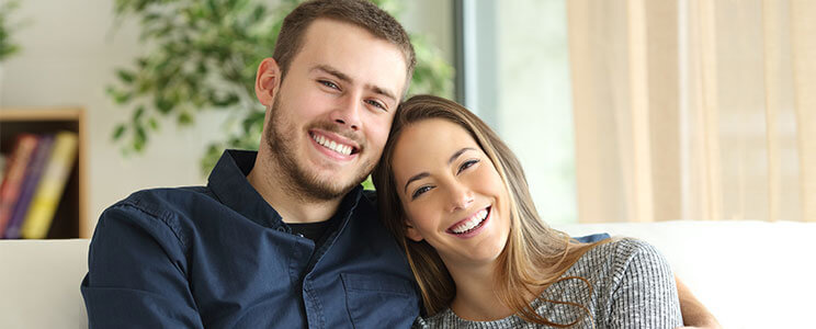 Photograph of a happy, smiling male & female couple holding each other while sitting on a couch in a home setting.